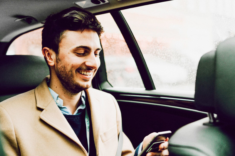 A happy man smiles and looks at his phone in the backseat of a car 800