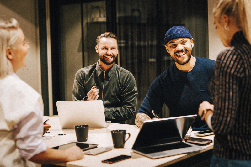 Four happy colleagues sit around a table with their computers and phones in the office 800