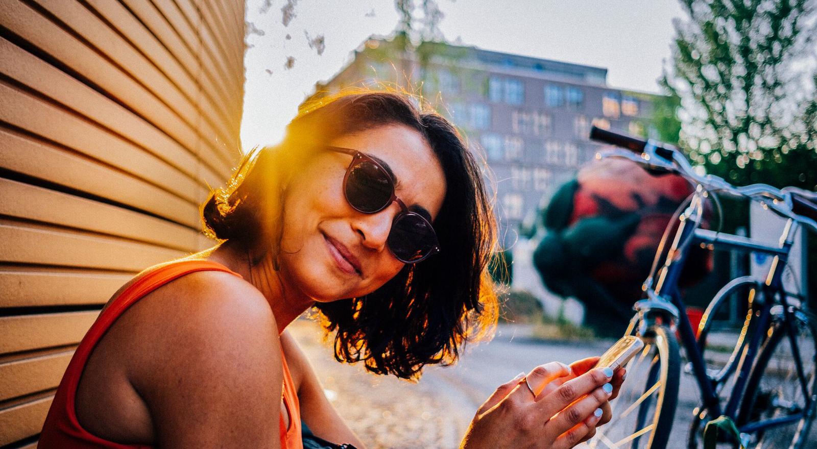 Large Web and Screen A happy woman wearing sunglasses rests and sits on the ground with her phone next to her bike outside