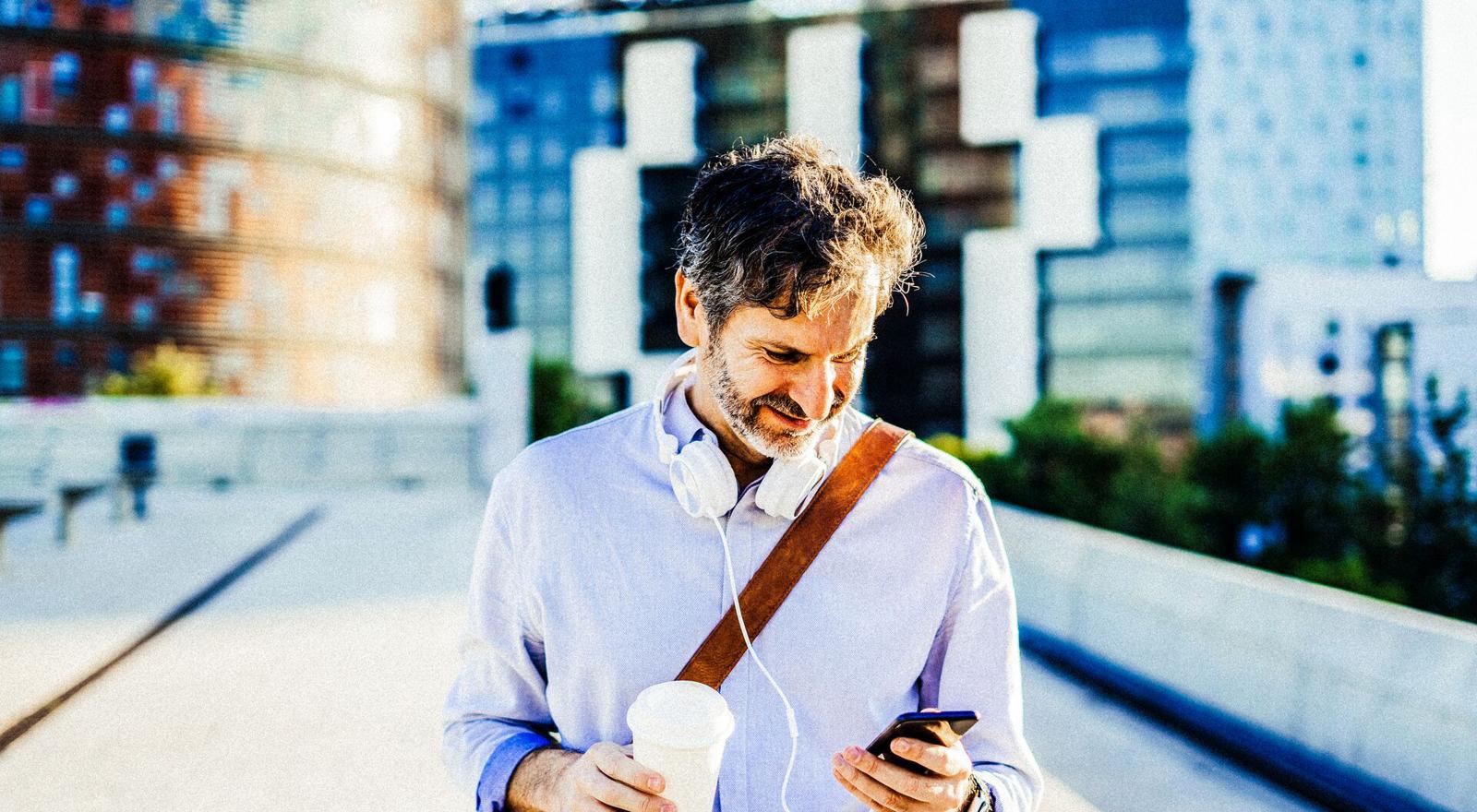 Mid adult man holding coffee cup looking at mobile phone with headphones around neck