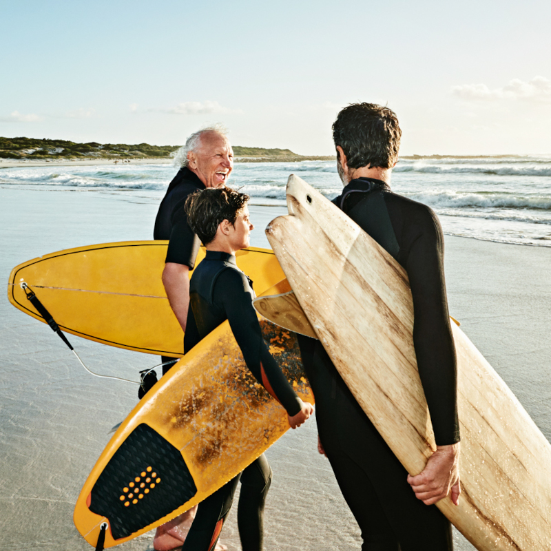 Three people in wetsuits stand in the sea with their surfboards 800x800